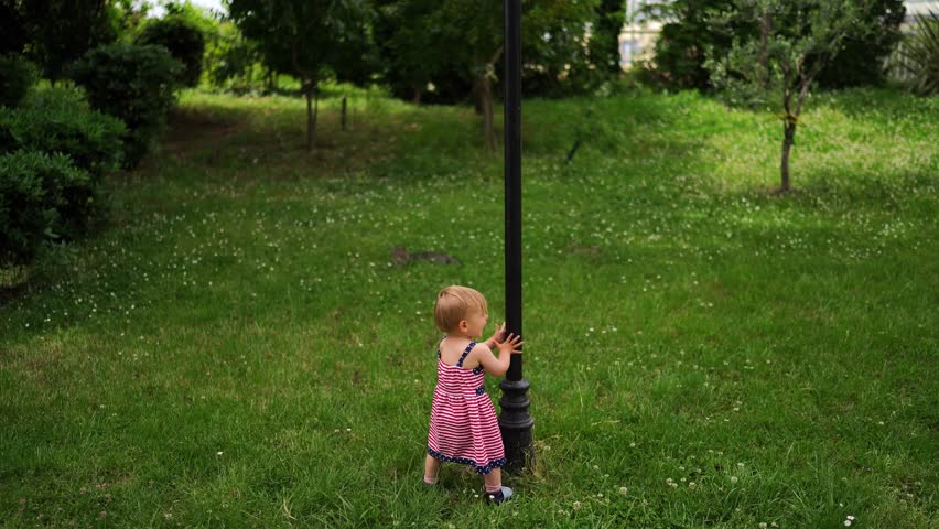 Little girl stands on a green lawn and waves her hands in front of a post