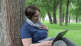 Senior woman having online conversation. Sitting on the grass in city park.