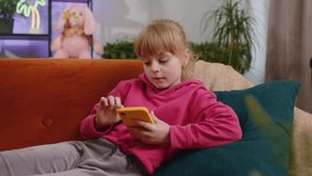 Young child kid girl texting messages on smartphone social media applications, watching relax movie, online shopping, play game. Female teen toddler uses mobile phone at home in room lying on sofa