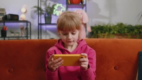 Worried funny child kid girl enthusiastically playing online racing or shooter video games on smartphone. Female teen toddler using smartphone gadget app with drive simulator at home sits on sofa