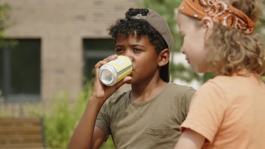 Medium close-up shot of two diverse young children in casual t-shirts and baseball caps hanging out together in the park, drinking soft beverages from cans, and chatting Royalty-Free Stock Footage #1101654977
