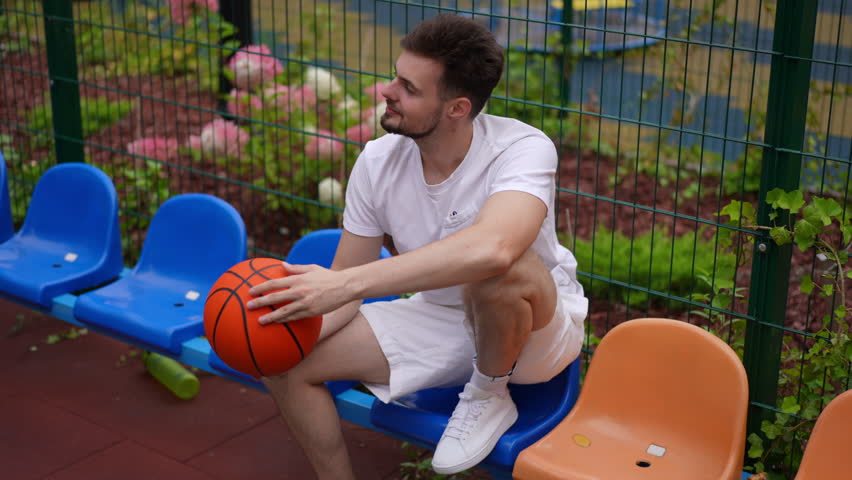 High angle view zoom in portrait of Caucasian sportsman smiling sitting with basketball ball outdoors turning looking at camera. Young fit man posing in slow motion with sport equipment