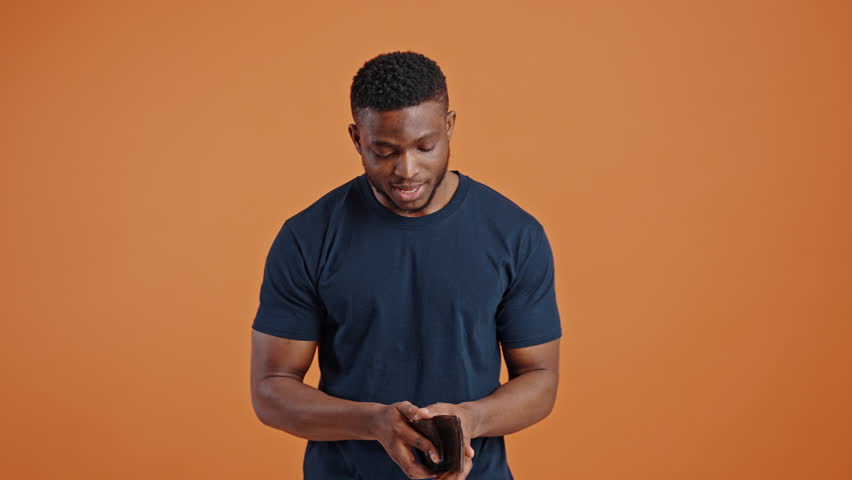 Young active dark-skinned man expressing sudden shock and confusion, throwing his leather purse down on the floor while find it empty, orange background. Spender people. Financial crisis. Unemployment Royalty-Free Stock Footage #1101657955