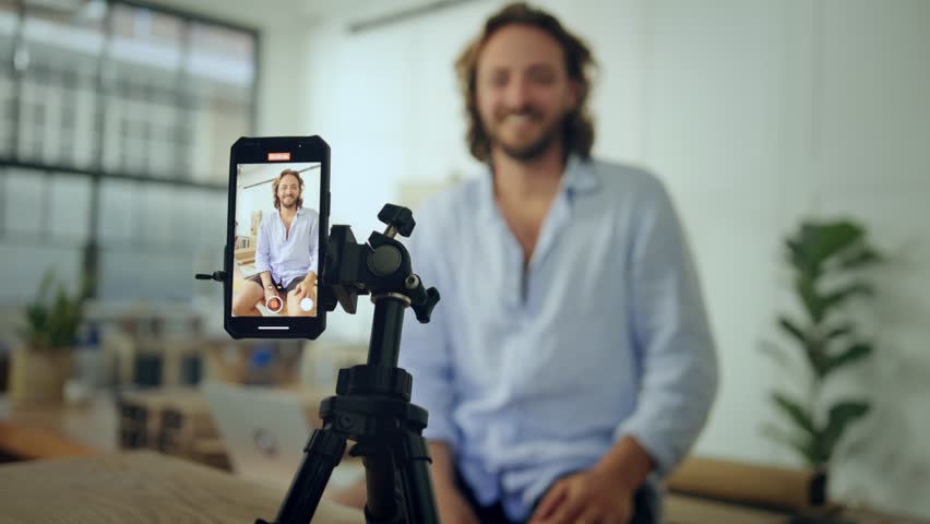Young caucasian man in shirt gesturing while sitting and talking on video by mobile phone in office