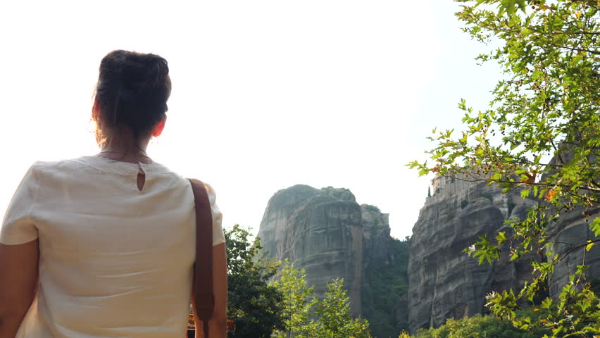 Young woman walking barefoot on a wall at Meteora rock mountains near Kalambaka, Greece. Large hat, sunglasses, fashion white dress and yellow skirt.
