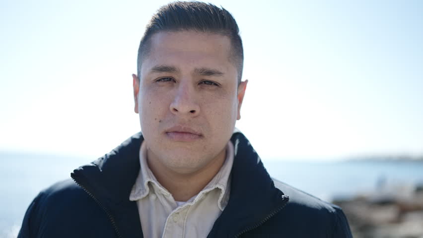 Young hispanic man standing with serious expression at seaside