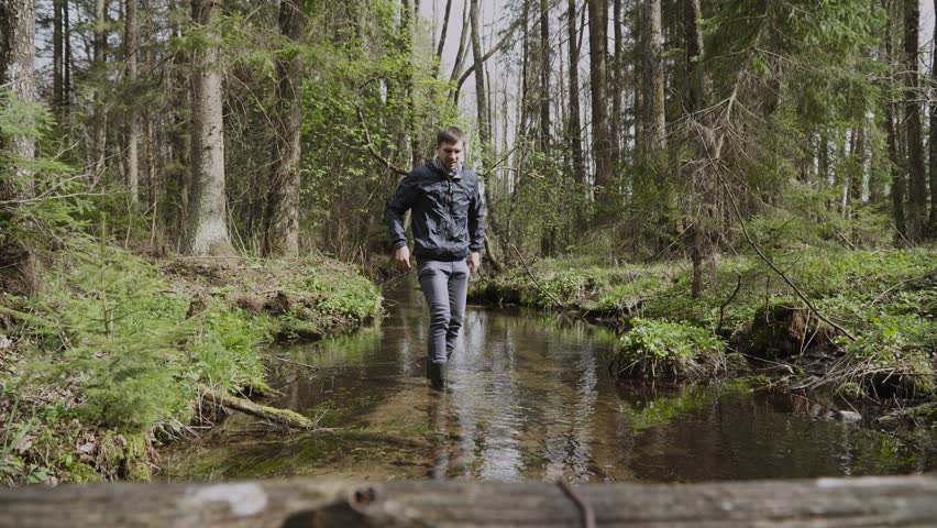 Thin man washes face with pure cold water in forest stream. Person in rubber boots sits down in bed of shallow clean creek, shakes off his wet hands. Bright spring sun, hard light shines on nature.