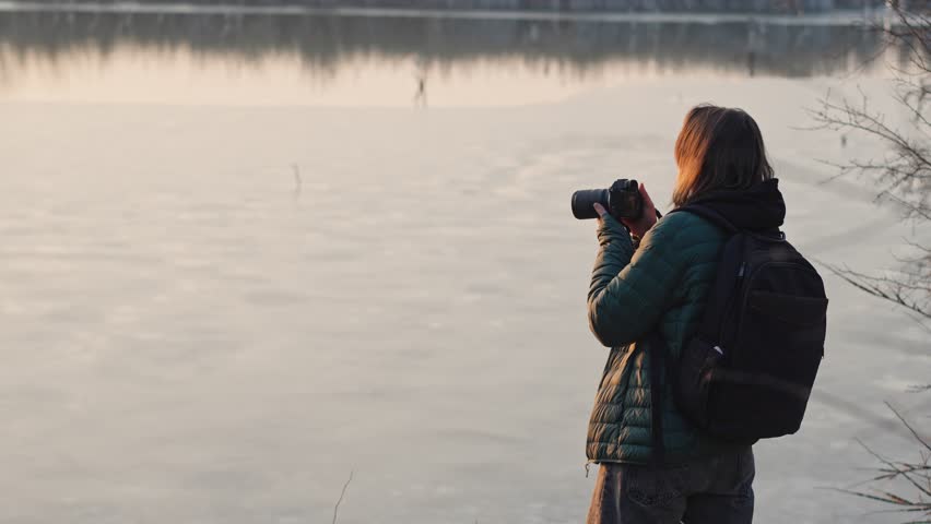 Slow-motion cinematic shot. A woman professional photographer taking a picture of a landscape while standing by the lake at the sunset time