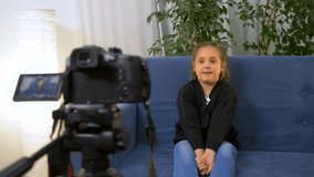 Happy little blogger girl live streaming video while talking to camera. little girl talks to camera while sitting on sofa in room. Child blogger sits at home speaking to audience live.