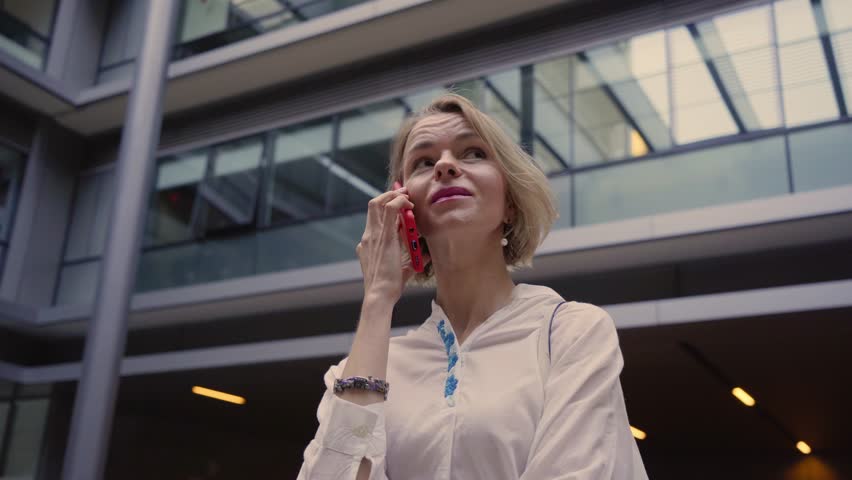 Smiling middle-aged blonde European woman secretary talking on cell outside her workplace. Lawyer answering mobile call in the hall of her business center. Professional people on the phone.
