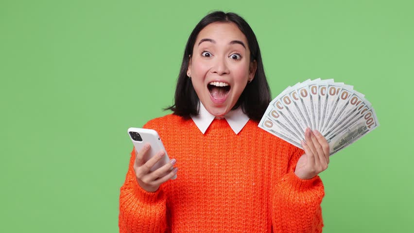 Excited happy young woman of Asian ethnicity she wear orange sweater use mobile cell phone hold win fan of cash money in dollar banknotes isolated on plain pastel light green color background studio