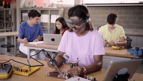 Looking at the camera video of a young Latina student with safety glasses in technical vocational school, lesson in High School. People soldering an electronic board.