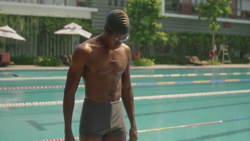 Medium long shot of athletic African American swimmer in cap, goggles and trunks going down pool stairs into water while training outdoors on summer day
