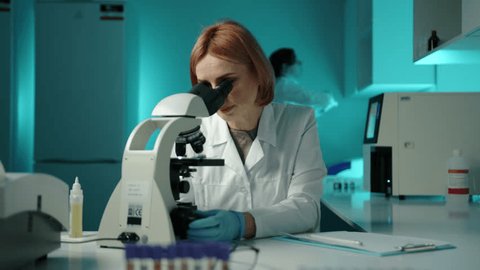 A woman in a white lab coat writing down something down and looking through a microscope. She is in a laboratory and is wearing glasses High quality 4k footage  库存视频