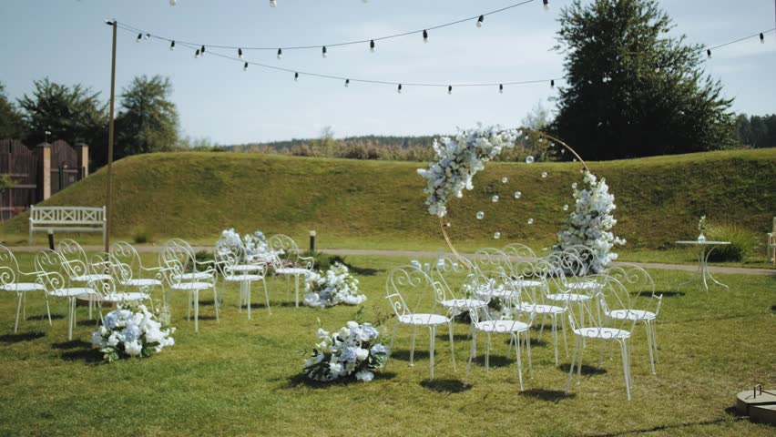 Side view of wedding arch decorations of flowers in white and blue colors with white metal chairs and lamps slow motion, outside wedding ceremony.