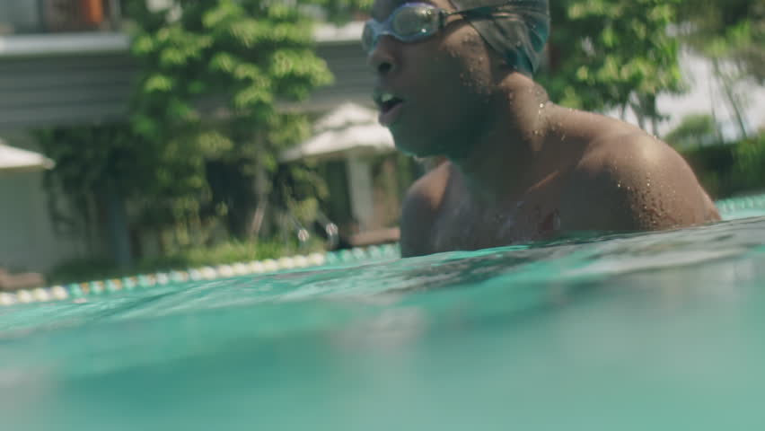 African American male athlete in goggles and cap diving into water and swimming freestyle in outdoor pool