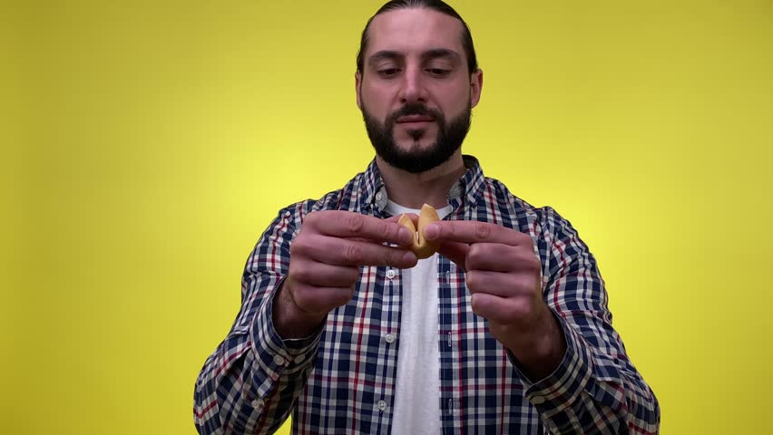 Mid adult bearded man opening fortune cookie, reading prediction, surprising good news isolated on yellow background. Positive male person wearing casual clothing. Luck concept, future forecasting