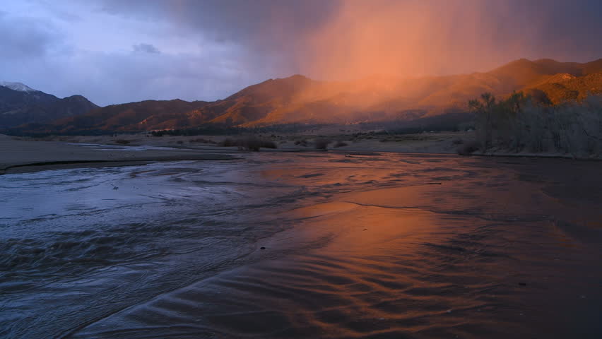 Sunset Medano Creek - A ray of twilight illuminates misty clouds hovering over rolling hills and rushing Medano Creek on a stormy Spring evening. Great Sand Dunes National Park, Colorado, USA.
