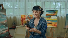 Happy smiling artist woman sitting on the chair at the art studio and holding her smartphone watching something