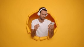 Young guy in white T-shirt posing in the middle of yellow backdrop, wearing headphones, smiling and dancing happily while listening music, lifestyle concept, copy space, high quality video