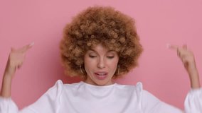 Portrait of young happy beautiful female with an afro hairstyle in a crisp white blouse against a pink studio background, she smiles and points to copy space down the screen, high quality video