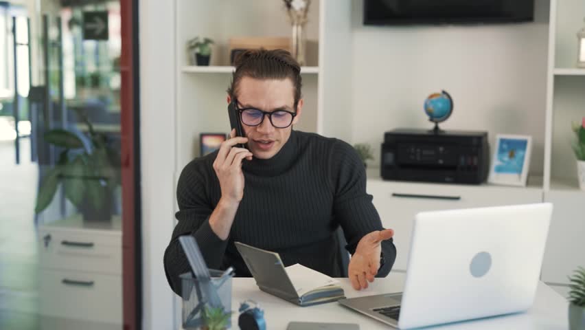 A man in glasses sits at a table and talks on the phone, occasionally laughing and enjoying the conversation.
