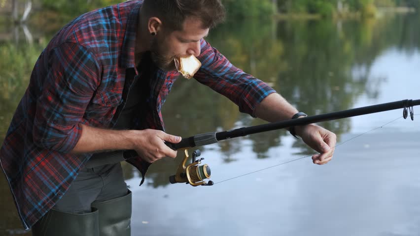 a fisherman adjusts a fishing rod while standing in the water and holding a sandwich in his mouth