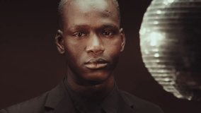 Conceptual close-up of emotionless Black man wearing elegant suit looking at camera, disco ball rotating on background - Powered by Shutterstock - Get 15% off with code: PIKWIZARD15