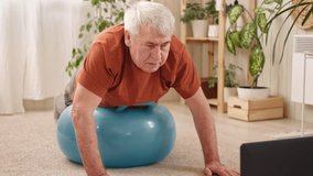 Mature senior man doing fitness exercises, gymnastics at home. Online lesson with laptop pc. Talking trainer. Recreation, well being. Elderly male exercising training, stretching. Old man working out