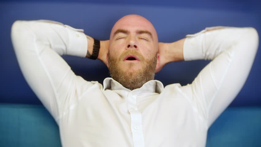 A man leaning back and relaxing at home on a sofa couch. The bald male has a beard and is wearing a white shirt.