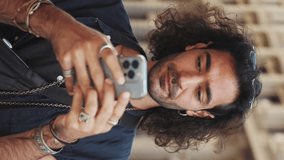 VERTICAL VIDEO: Young attractive italian guy with long curly hair and stubble is using mobile phone. Stylish man determines the location using map on his smartphone
