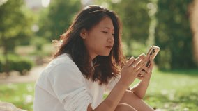 Young Asian woman with long brown hair wearing white t-shirt sitting in park having picnic on summer day outdoors, using cell phone. Girl browsing social networks, photo, video on smartphone