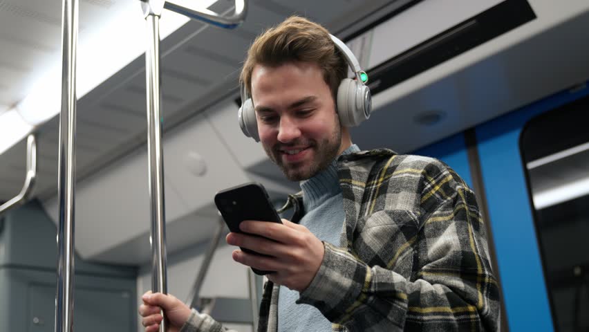 Cheerful man in wireless headphones rides the subway, uses a smartphone and surfs the internet, a student goes to study at the university, morning on the public transport.