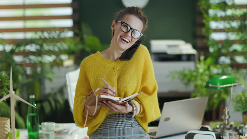 happy trendy small business owner woman in yellow sweater with notebook speaking on a smartphone in the modern green office. Royalty-Free Stock Footage #1101707343