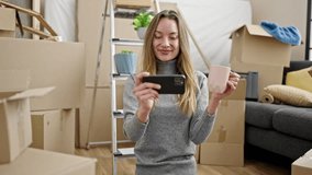 Young caucasian woman sitting on the floor drinking a cup of coffee watching video on smartphone at new home