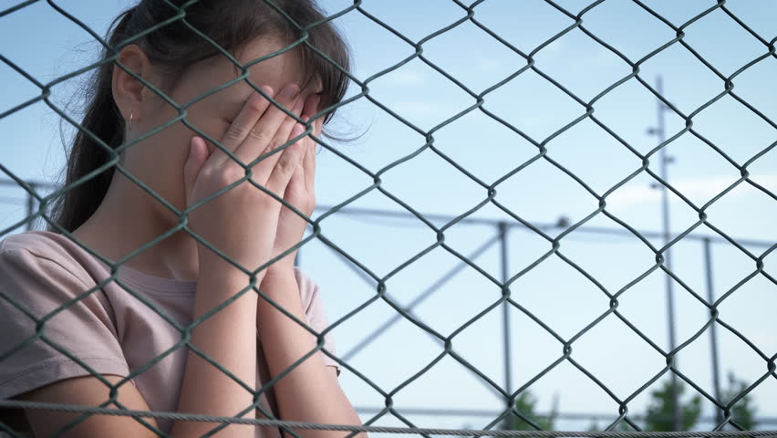 A crying teenager in a refugee camp. A teenage girl hides her face with her hands behind a wire fence.