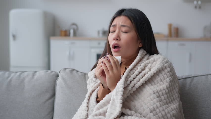 Unhealthy ill sick Asian woman multiethnic korean chinese girl patient covered in warm blanket at home blowing to paper napkin sneeze runny nose allergy flu caught cold sneezing has covid-19 symptom Royalty-Free Stock Footage #1101721329