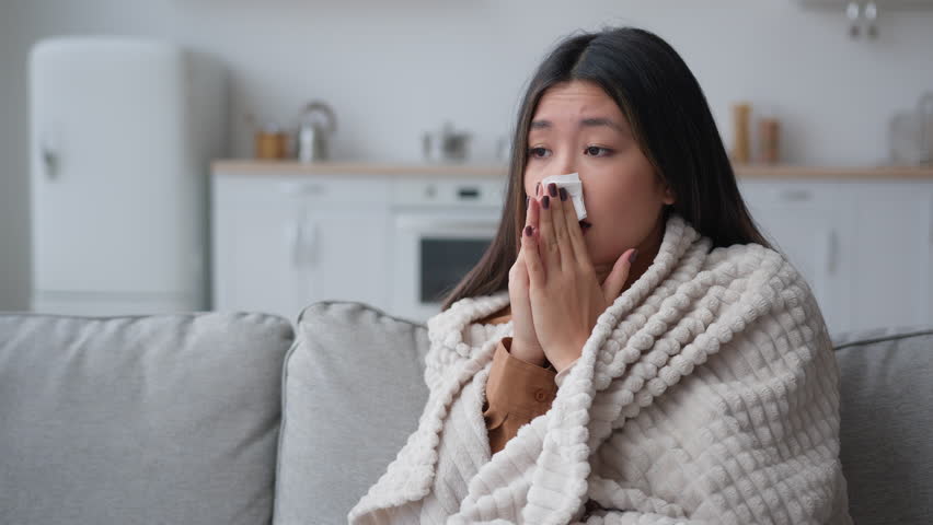Unhealthy ill sick Asian woman multiethnic korean chinese girl patient covered in warm blanket at home blowing to paper napkin sneeze runny nose allergy flu caught cold sneezing has covid-19 symptom