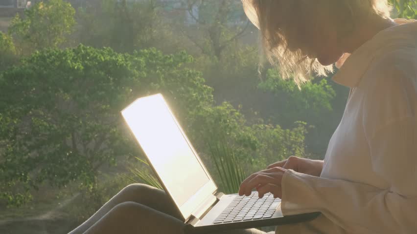 Close-up of young woman typing on a laptop keyboard while sitting on the rooftop with a beautiful green garden view at the sunrise morning time. | Shutterstock HD Video #1101734009