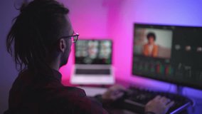 Man video editor wearing headphones sits at computer with backlight, looks in monitor. Video editing with music and color correction. Freelance, working in home office . Professional video processing.