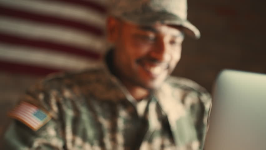 Man in camouflage clothing video calls his loved ones with a laptop, with an American flag in the background. Patriotic soldier maintaining his long distance relationship with his family during deploy Royalty-Free Stock Footage #1101736211