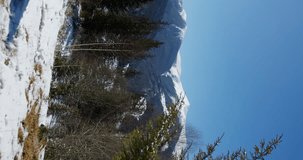 a tourist girl in a bright jacket on a sunny day enters the frame against the background of snow-capped mountain peaks and covers her eyes from the sun's rays with her palm. vertical video