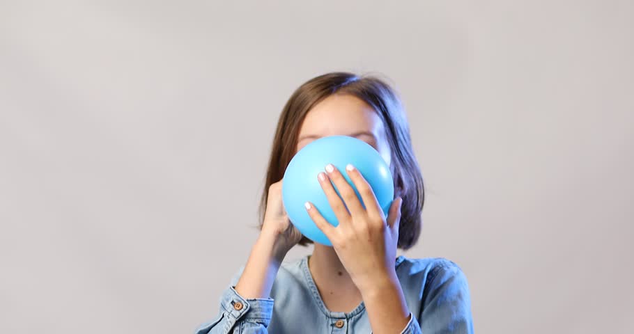 Pretty little girl in casual denim dress blowing, inflate blue balloon on white background, studio