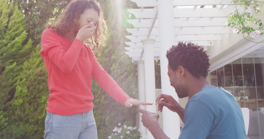 Happy biracial man proposing on knees with ring to surprised woman. romance and tradition, marriage proposal and engagement ring.