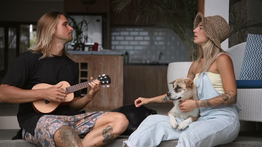 Happy family love sing fun song. Talent person play small ukulele guitar. Pet lovers people. Nice couple enjoy date. Guy rub cute pooch dog. Joyful girl rest yard. Two doggy hound sit porch. Pets care Royalty-Free Stock Footage #1101745323