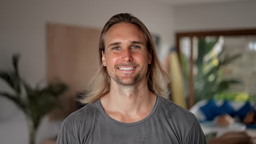 Happy male person face home portrait. Blue eyes look at camera close up. Joyful bristle surfer man long hair. Smiling shy guy. Kind people portraits. Young adult hippie inside house. Hipster hairstyle