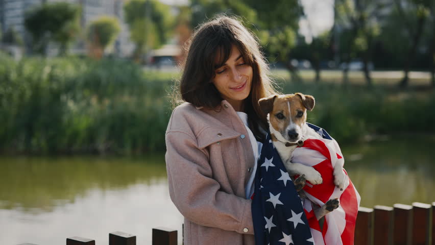 Portrait of the Caucasian Woman Owner Smiling Holding Her Little Jack Russell Wrapped in the USA Flag. Girl Standing Outside in the Park Looking at the Camera. People and Dog Patriotism Concept | Shutterstock HD Video #1101745485