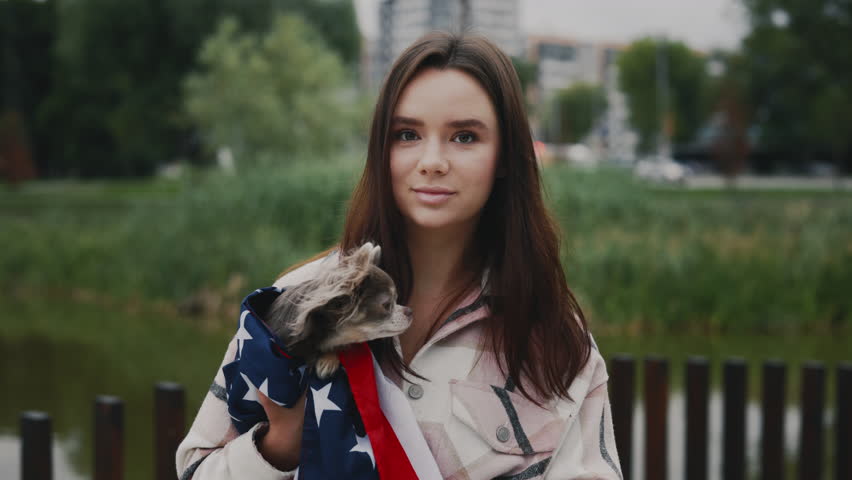 Portrait of the caucasian smiled woman owner smiling holding her small chihuahua wrapped in the usa flag standing outside in the park looking at the camera. People and dog patriotism concept | Shutterstock HD Video #1101745497
