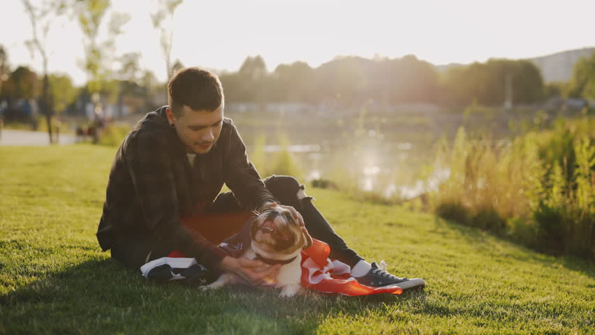 Handsome Smiled Stylish Man Sitting on the Grass Playing with Brooklyn Bulldog Wrapped in the USA Flag, Sitting Near His in the Park. Patriotic Dog Is Wrapped in the American Flag | Shutterstock HD Video #1101745499