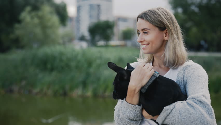 The Happy Caucasian Pet Owner is Stroking and Kissing Her Black French Bulldog Puppy, Showing Love to the Dog Standing in the Park near Local Lake. People and Dog Friendship Concept | Shutterstock HD Video #1101745513